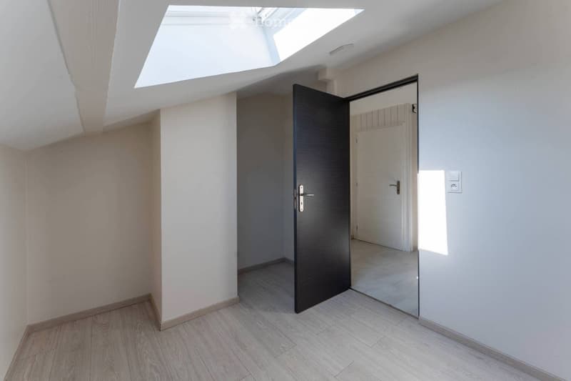 Bright, empty hallway with wooden flooring, white walls, and a skylight above. A dark wooden door leads to another room.