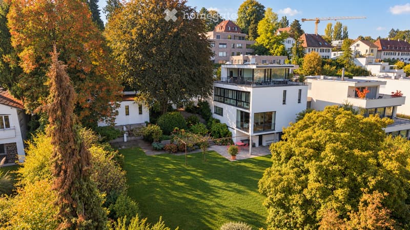 Aerial view of a modern residential complex with multiple white buildings, surrounded by lush greenery and trees. The central building features large windows and a balcony, while the surrounding area includes well-maintained lawns and gardens.
