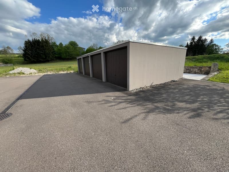 A modern, single-story garage with three large roll-up doors, situated on a paved driveway with a stone wall and greenery in the background.