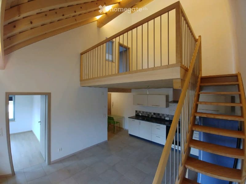 Open hallway with wooden staircase leading to an upper level, kitchen visible to the right with white cabinets, black countertops, and a green chair.