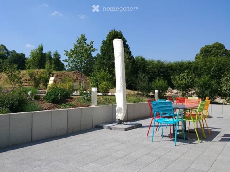 A modern outdoor seating area with colorful chairs and a table, surrounded by a low concrete wall and lush greenery.