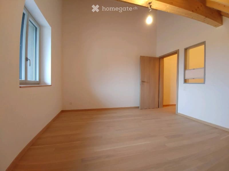Empty room with wooden flooring, white walls, and exposed wooden beams on the ceiling. Features two windows and an open doorway leading to another space.