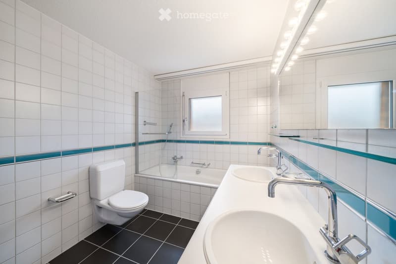 Modern bathroom with white tiles, black floor tiles, a toilet, a bathtub, a double sink with chrome faucets, and a large mirror with built-in lighting.