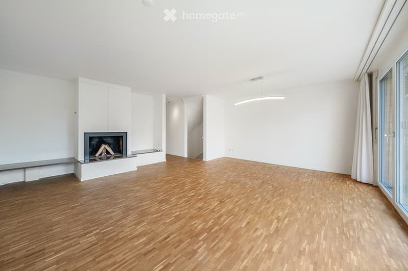 Bright, unfurnished room with wooden flooring, white walls, and a built-in fireplace with a black frame and a small bench. A large window with white curtains is on the right side, and a modern pendant light hangs from the ceiling.