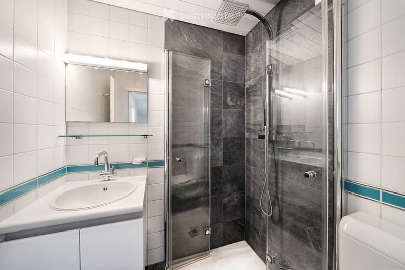 Modern bathroom with white tiles, a glass-enclosed shower, a white sink with a chrome faucet, a mirror, and a glass shelf above the sink.