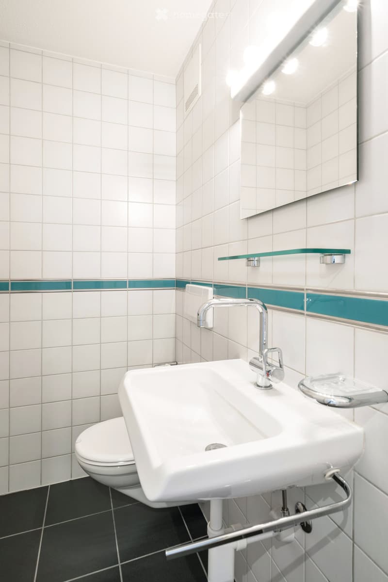 Modern bathroom with white tiles, a white sink, a chrome faucet, a toilet, and a large mirror above the sink. The walls feature a decorative teal accent strip.