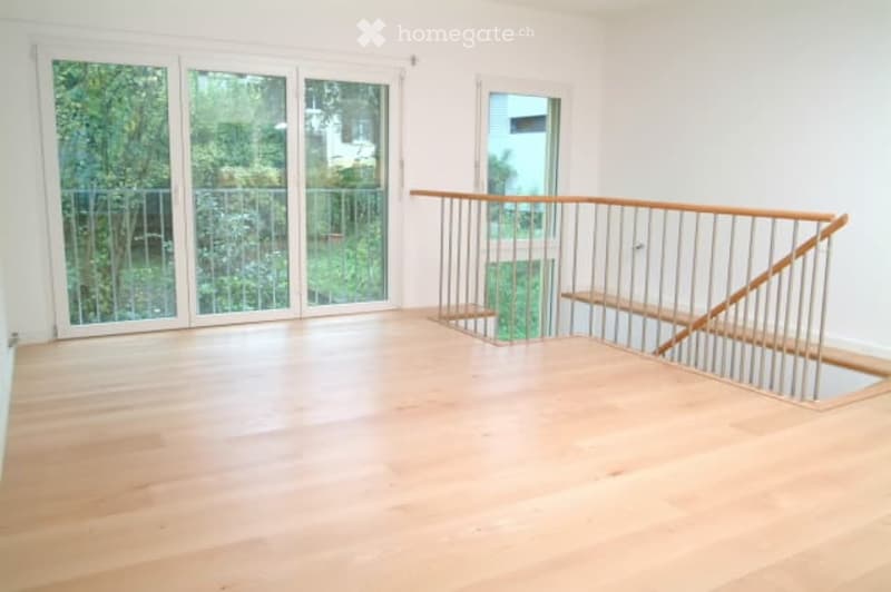 Empty room with wooden flooring, large glass sliding doors leading to a balcony, and a modern metal staircase railing.
