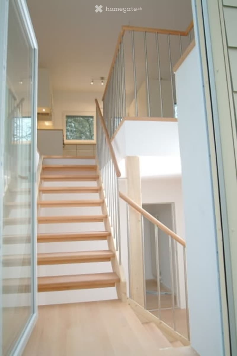 Modern interior staircase with wooden steps, glass railings, and white walls.