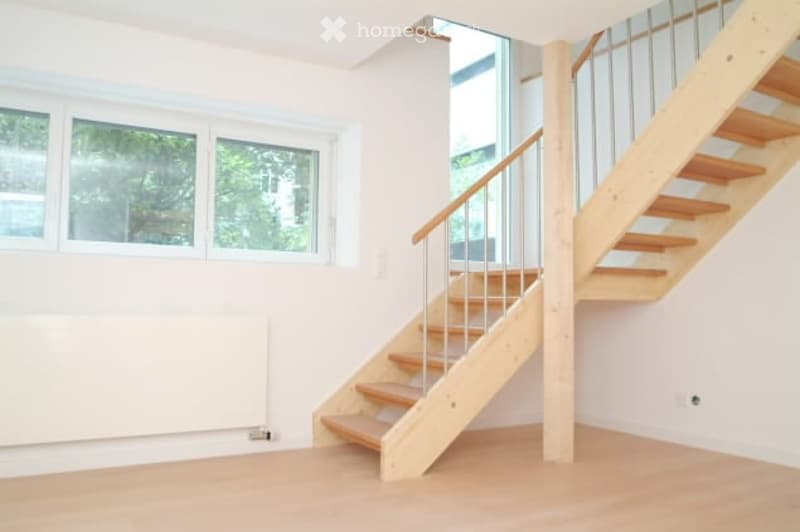 Empty room with wooden floor, white walls, large windows, and a staircase leading to an upper level.
