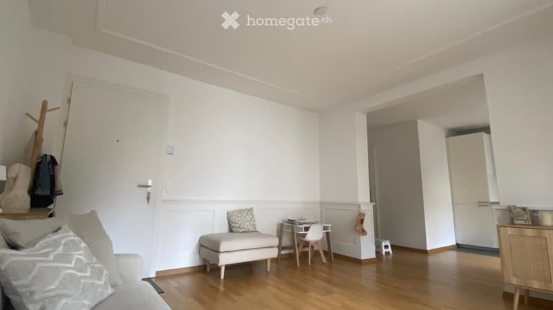 A modern, well-lit living room with a grey sofa, a small wooden table with a chair, a white stool, and a coat rack near the door. The room features light-colored walls, wooden flooring, and a small cabinet in the corner.
