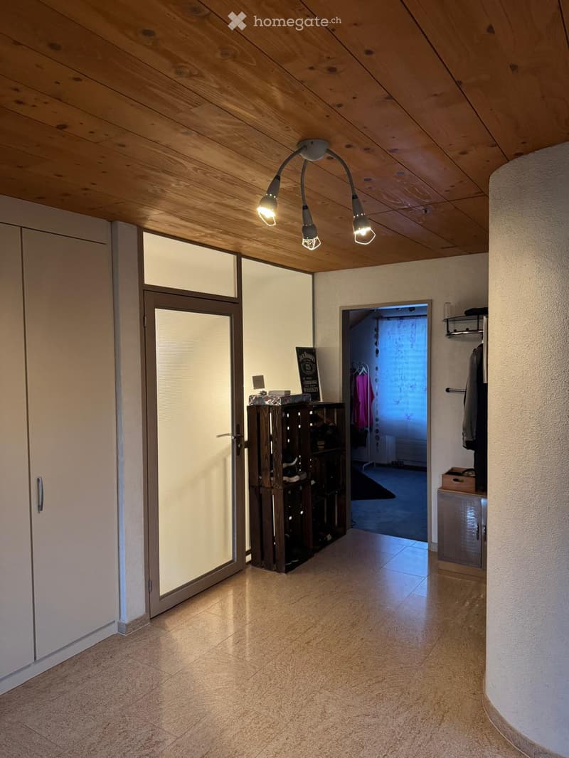 Bright hallway with wooden ceiling, tiled floor, and modern lighting fixture. Features a glass door, built-in shelving unit, and a coat rack with hanging clothes.