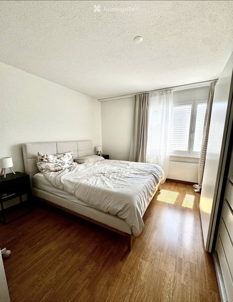 A well-lit bedroom with wooden flooring, a large bed with a headboard, nightstands with lamps, and a window with curtains.