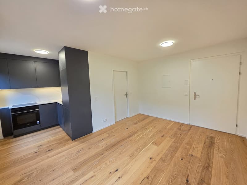 Empty room with wooden flooring, white walls, and a small kitchen area with dark cabinets and a built-in oven. Two white doors are visible, one leading to another room and the other to an exit.