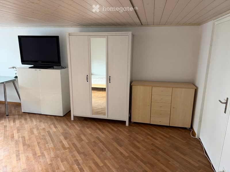 A furnished room with a television on a cabinet, a large mirrored wardrobe, a wooden cabinet with drawers, and a glass table with metal legs.