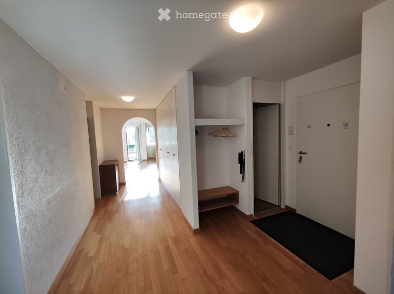 The image shows the interior of an apartment. The hallway has hardwood floors and white walls. There is a closet with a hanging rod and shelves. The hallway leads to other rooms, which are not visible in the image. A washing machine is visible, indicating