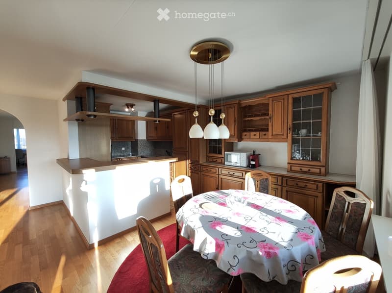 The image shows a combined kitchen and dining room area. The kitchen features wooden cabinets and drawers, a countertop, and a dishwasher. The dining area has a round table with a patterned tablecloth and chairs. The room is well-lit with pendant lights a