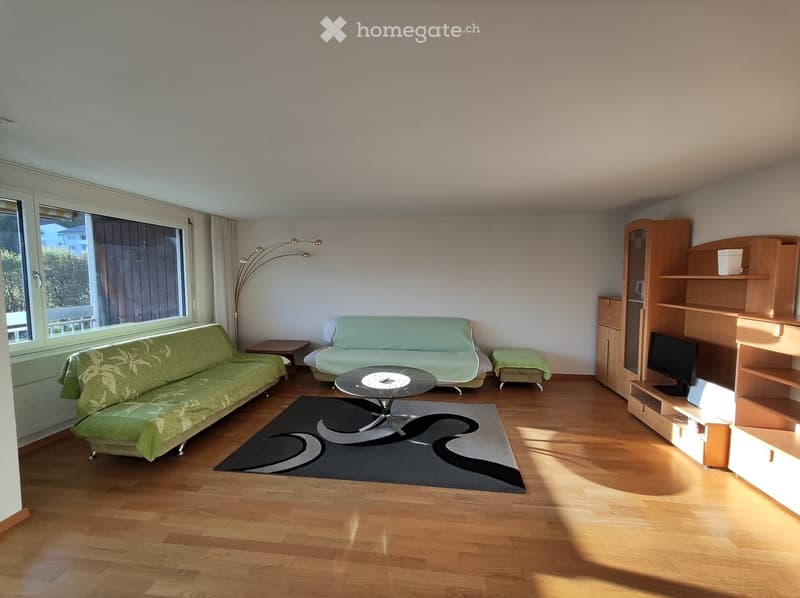 Spacious living room with large windows, hardwood floors, green patterned sofa, coffee table, and a wall unit with a TV and shelves.