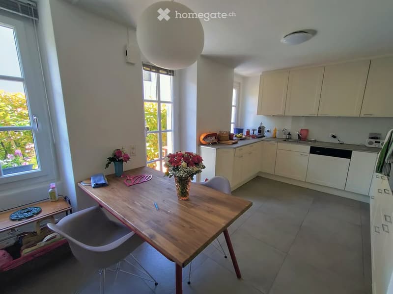 Bright kitchen with light-colored cabinets, a wooden dining table with chairs, and a vase of flowers on the table. The kitchen features a sink, dishwasher, and various small appliances.
