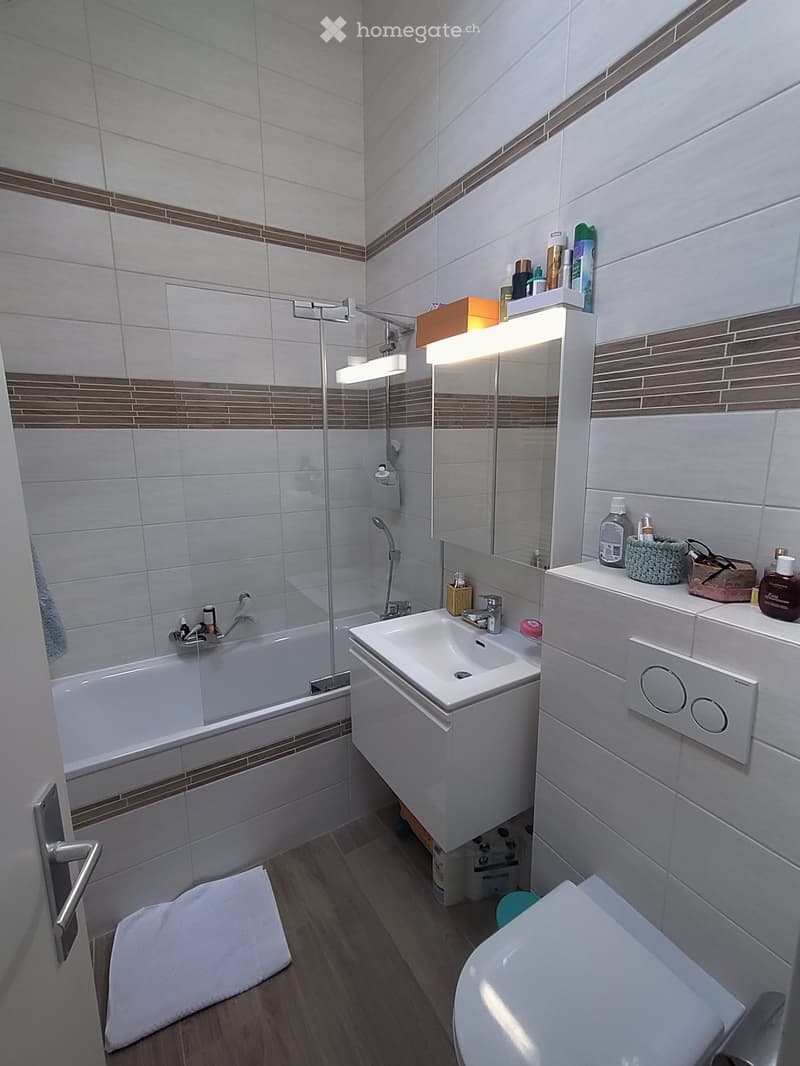Modern bathroom with white tiles, a bathtub, a sink, a toilet, a large mirror with built-in lighting, and a towel on the floor.