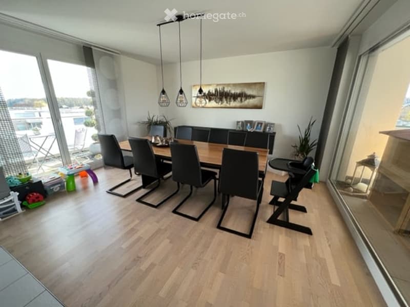Bright dining room with wooden floor, large windows offering a view, a dining table with six chairs, a baby chair, pendant lights, and a decorative picture frame on the wall.