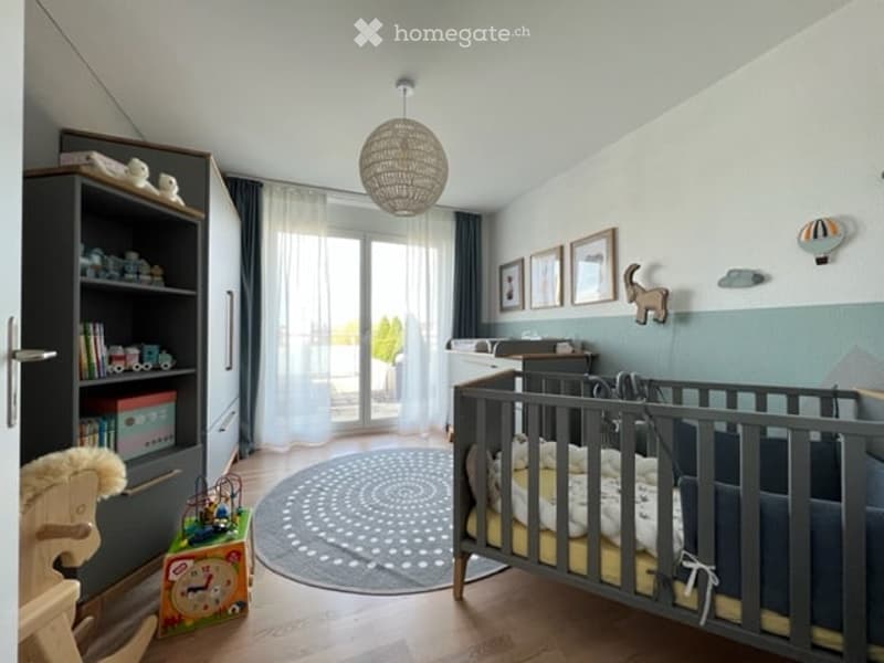 A well-lit, modern nursery with a crib, bookshelf, toys, and a large window with curtains. The room features a round rug, framed artwork, and a decorative balloon on the wall.