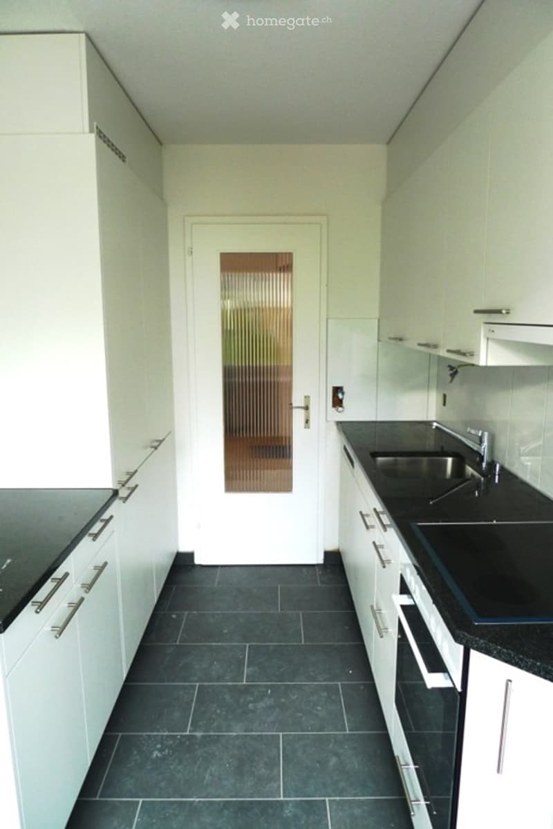 Modern kitchen with white cabinets, black countertops, and a stainless steel sink. The floor is dark tiled, and there is a door with a glass panel leading to another room.