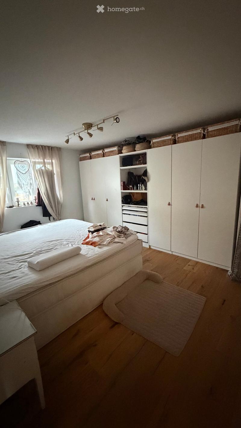 Bedroom with a wooden floor, a bed with white bedding, a wardrobe, and a window with curtains. There are baskets on top of the wardrobe and a small rug on the floor.