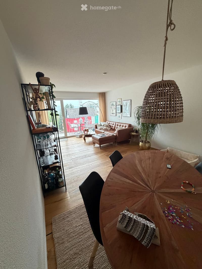 Bright living and dining area with wooden flooring, a hanging woven lamp, a leather sofa, a coffee table, a dining table with chairs, and a shelf with decorative items.