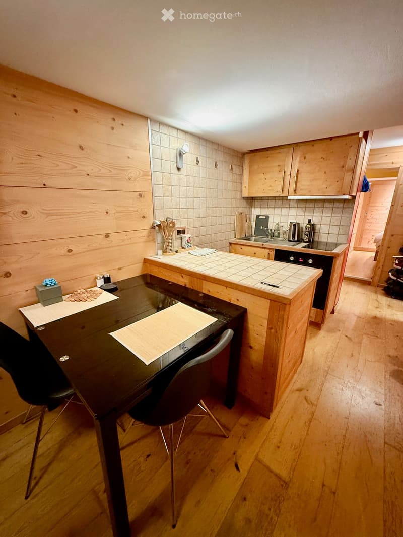 The kitchen features wooden cabinets, a black dining table with chairs, and a countertop with various kitchen items including a cup, bottle, and utensils.
