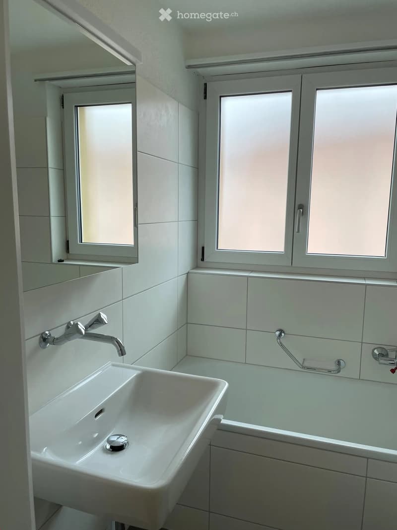 Modern bathroom with white tiled walls and floor, a white sink with a chrome faucet, and a bathtub with a chrome grab bar.