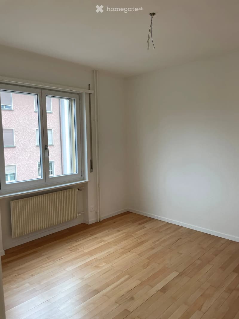 Empty room with wooden flooring, white walls, large window with radiator below, and visible electrical outlet.