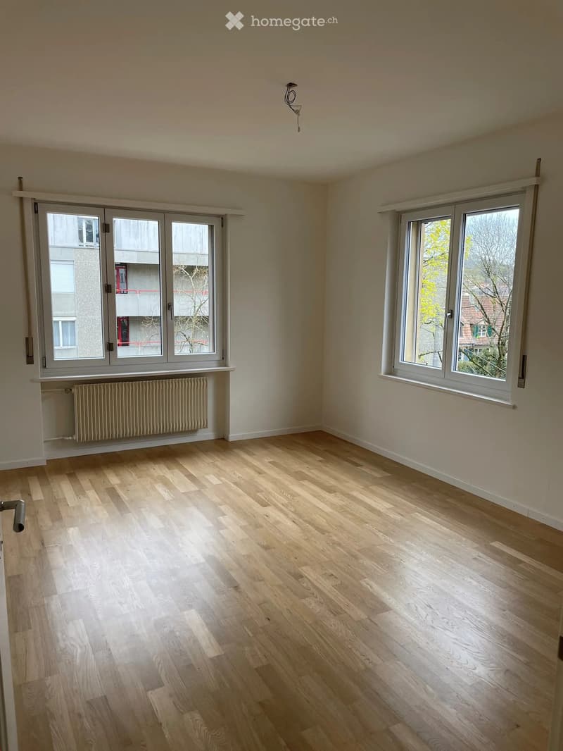 Empty room with light wooden flooring, white walls, large windows, and a radiator under one window.