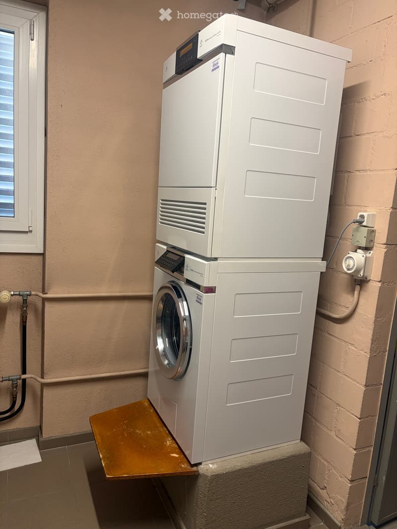 A compact bathroom with a stacked washing machine and dishwasher unit, beige walls, and a small wooden shelf.