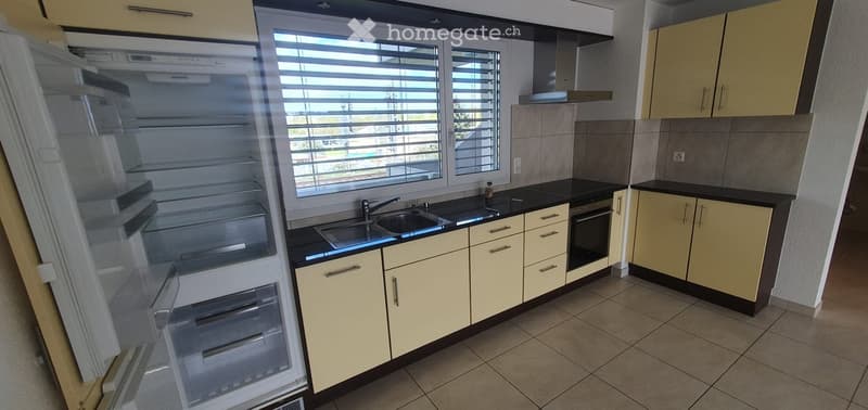 Modern kitchen with light yellow cabinets, black countertops, stainless steel sink, and an open refrigerator. The space features large windows with blinds and a tiled floor.