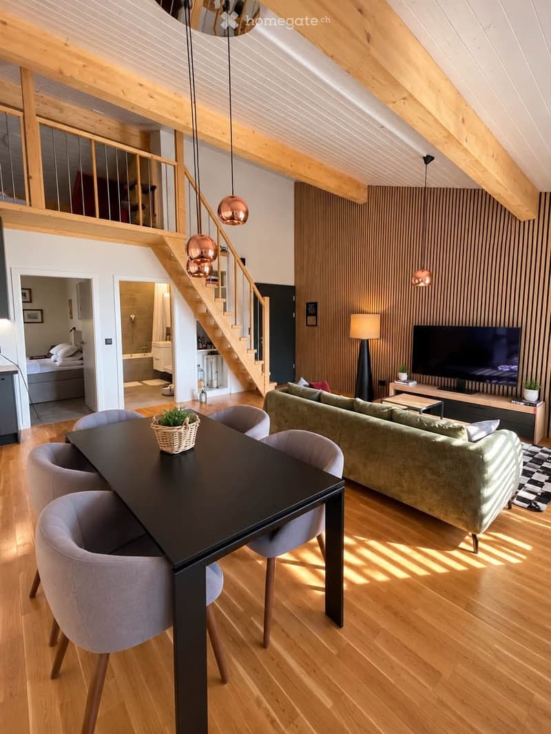 Modern living and dining area with wooden floors, exposed wooden beams, a green sofa, a black dining table with four chairs, a television on a wooden stand, and a staircase leading to an upper level.