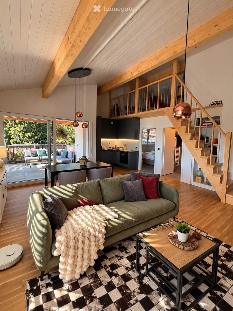 Open-plan living and dining area with a green sofa, coffee table, dining table, and chairs. The space features wooden floors, exposed wooden beams, pendant lights, and a staircase leading to an upper level. A modern kitchen is visible in the background, a