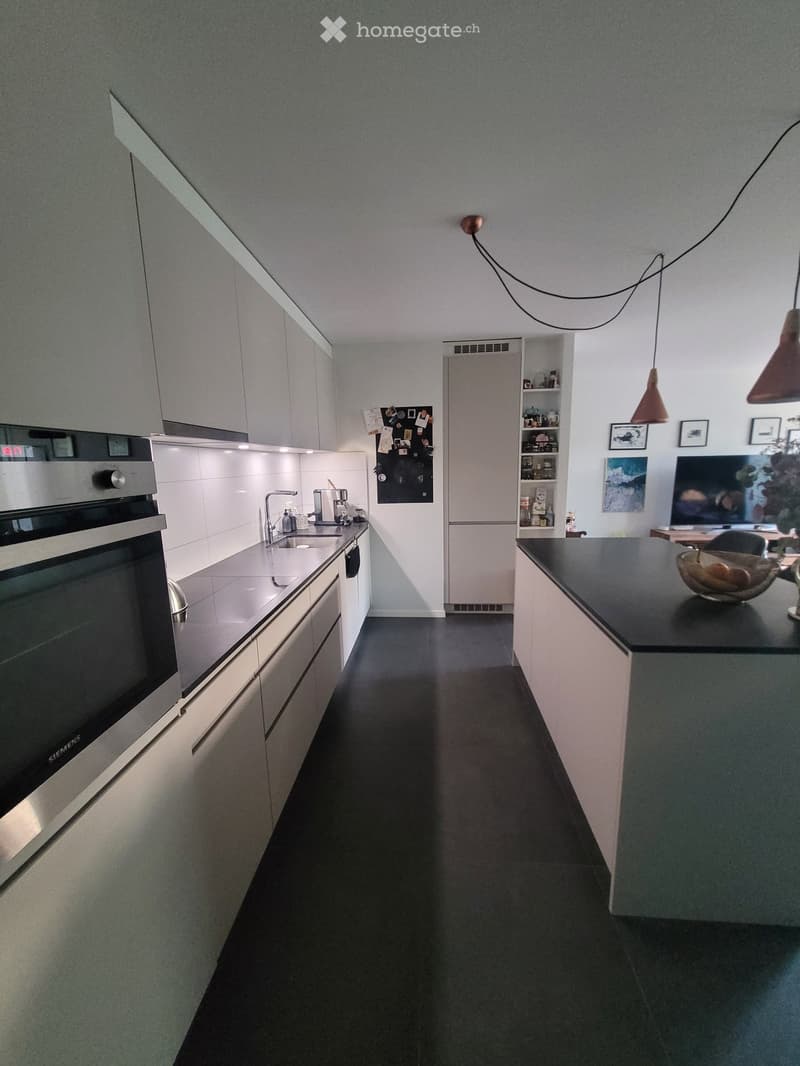 Modern kitchen with white cabinets, stainless steel oven, sink, and counter. Features pendant lights, a bowl on the counter, and framed pictures on the wall.