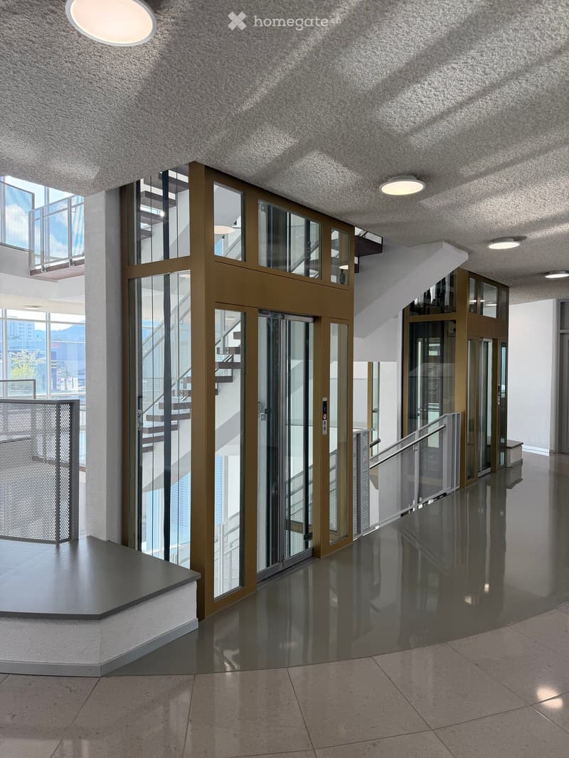 Modern hallway with polished tile floors, glass elevator doors, and recessed lighting. The area features a staircase with a metal railing and a view of the exterior through large windows.