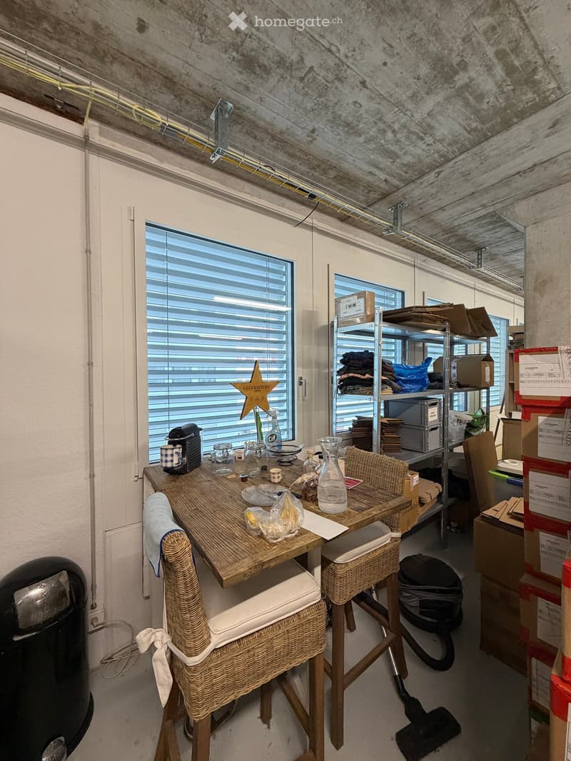 A small, furnished room with a wooden table, two wicker chairs, a coffee machine, and various items on the table, including cups, bottles, and a vase. Shelves with folded clothes and boxes are visible in the background.