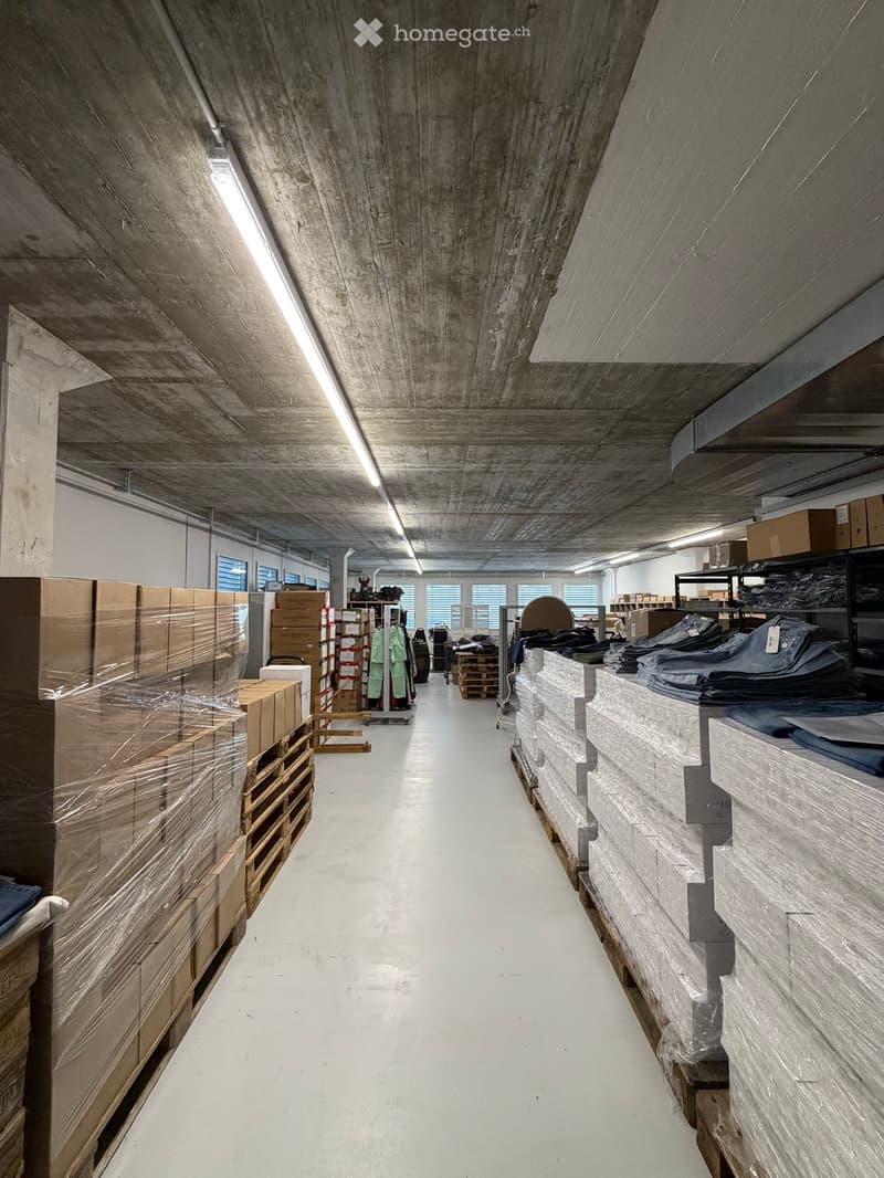 A long, empty corridor with exposed concrete and wooden ceiling beams, white flooring, and large windows at the far end. Stacks of boxes and pallets line both sides.