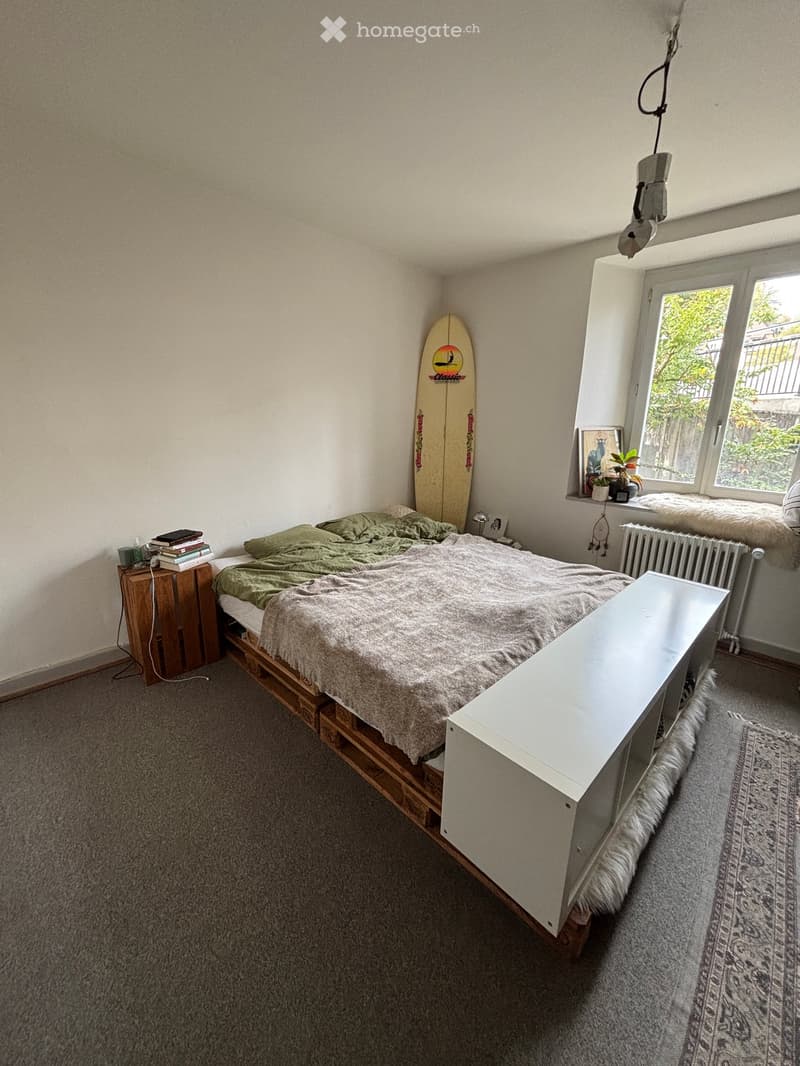 Cozy bedroom with a wooden bed frame, green bedding, nightstand, surfboard leaning against the wall, and a small table near the window.