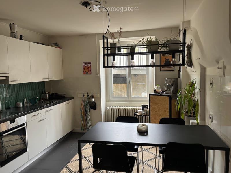 Modern kitchen with white cabinets, black countertops, a dining table with chairs, and a coffee maker on the counter. There is a window with a view, and several plants are placed around the room.