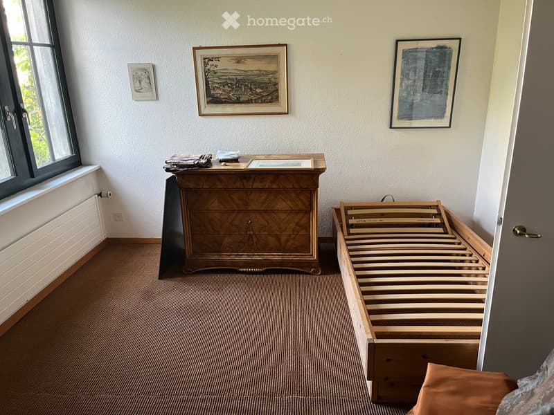 Bedroom with a wooden bed frame, a wooden chest of drawers, and several picture frames on the wall. The room has carpeted flooring and a window with a view of greenery.