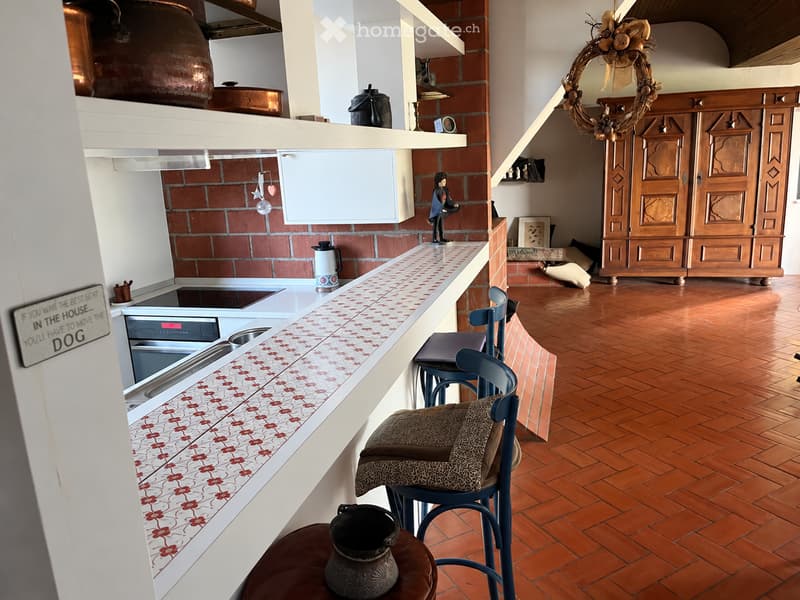 A cozy kitchen area with a brick wall, wooden floor, and a small seating area with a stool and chair. The kitchen features a stove, refrigerator, and a countertop with various items, including a teapot and a figurine. A wooden cabinet and a decorative wre