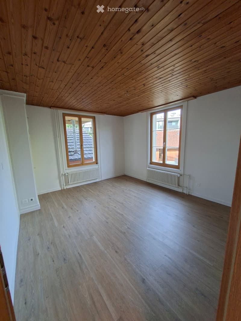 Empty room with wooden flooring, white walls, and large windows with wooden frames. Radiators are visible beneath the windows.