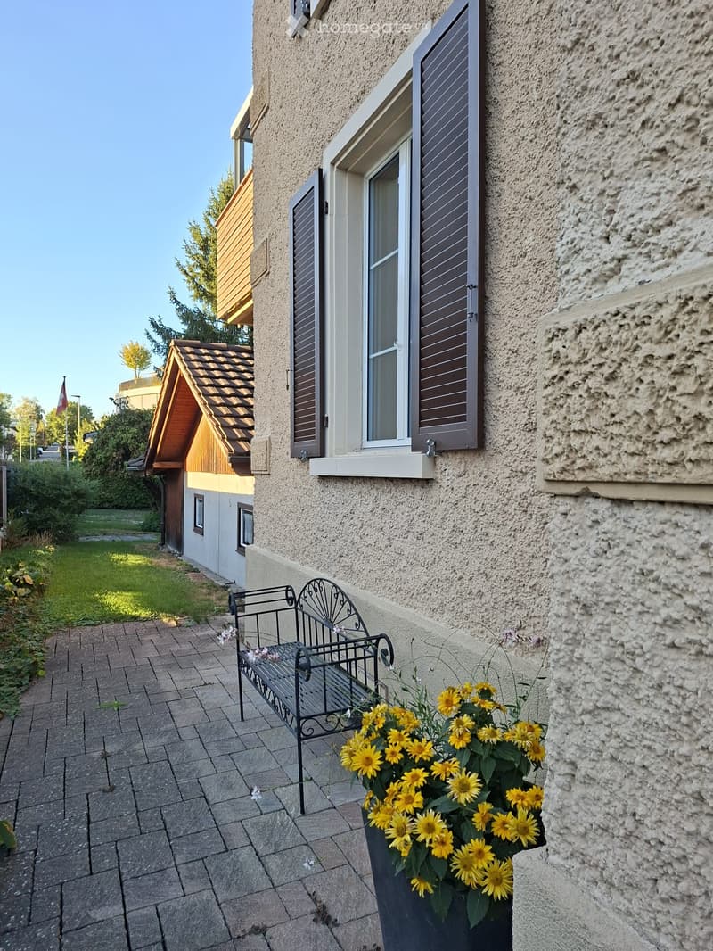 Balcony area with a black metal bench, a flower pot with yellow sunflowers, and a paved brick pathway leading to a garden.