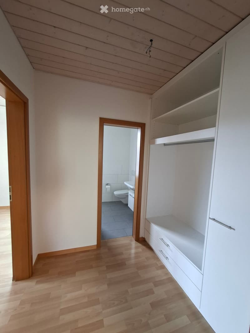 An unfurnished room with wooden flooring, white walls, and built-in white shelving units. A doorway leads to a small bathroom with a toilet, sink, and tiled floor.