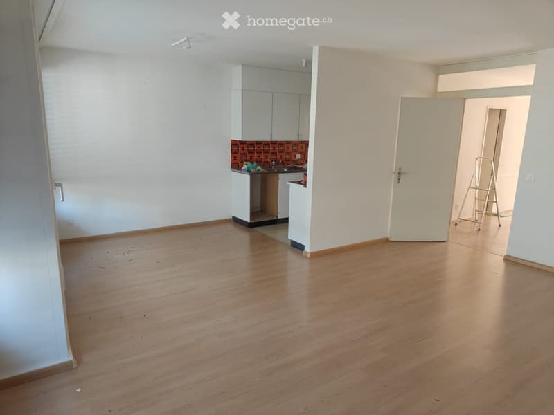 Empty room with wooden flooring, white walls, and a small kitchen area visible in the background.