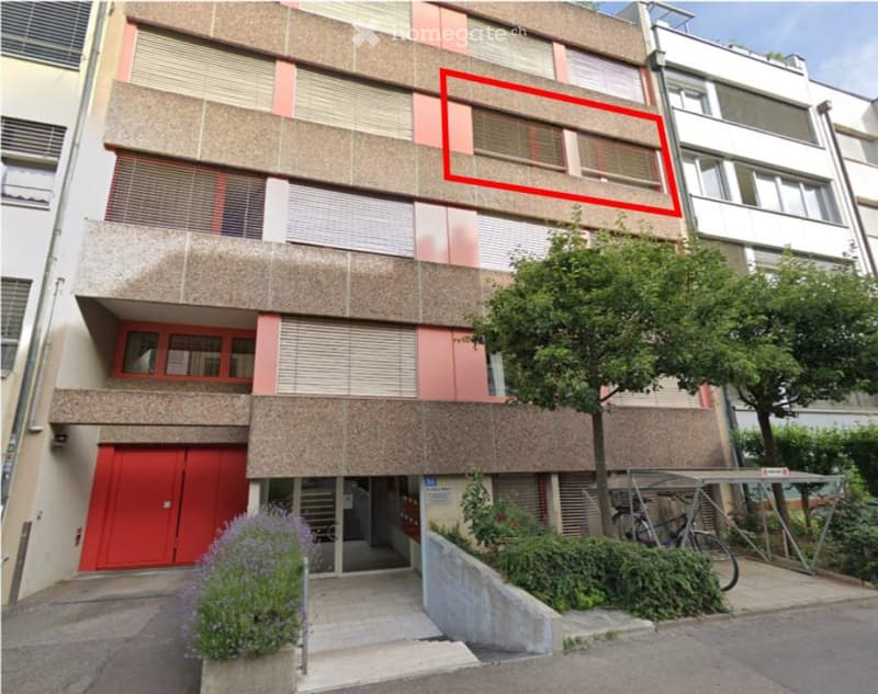 Multi-story apartment building with a red garage entrance, multiple windows, and a small garden area with bicycles and plants.