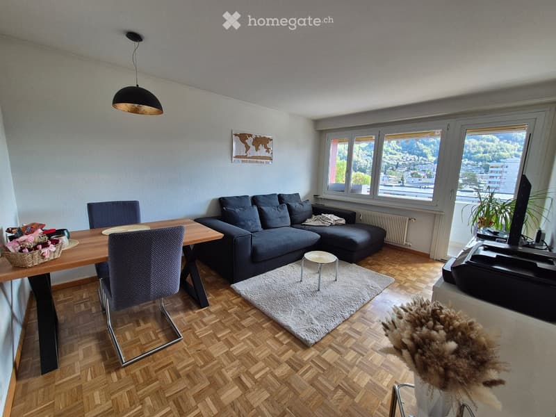 Living room with wooden flooring, large windows offering a scenic view, a grey sectional sofa, a coffee table, a small round stool, a dining table with chairs, a television, and a pendant light.